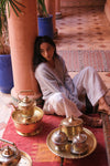 A woman in a light gray robe and white pants sits on a red floor mat in a Moroccan-style courtyard, surrounded by brass and silver tea sets and decorative columns.