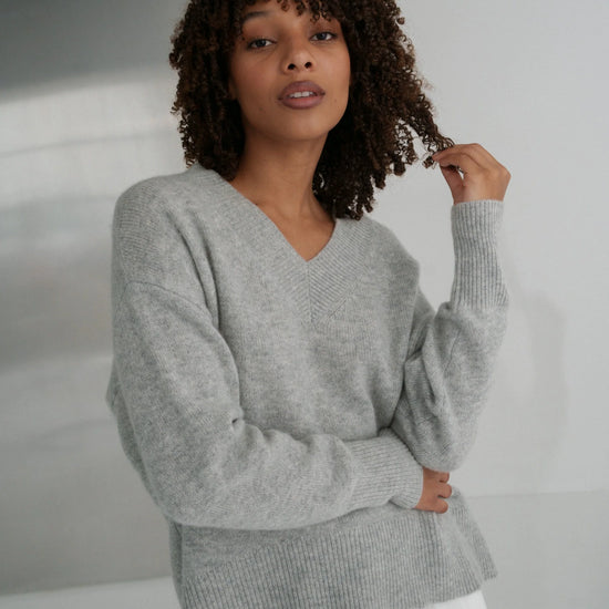 Woman wearing a gray sweater and white pants against a neutral background