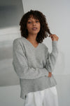 Woman wearing a gray sweater and white pants against a neutral background