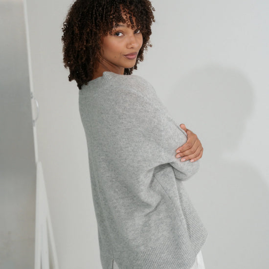 Woman wearing a gray sweater and white pants against a plain background