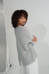 Woman wearing a gray sweater and white pants against a plain background