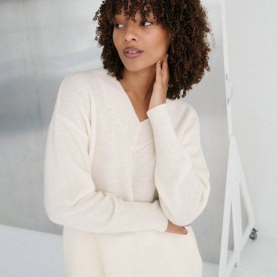 Woman wearing a cream sweater and white pants against a plain background
