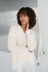 Woman wearing a cream sweater and white pants against a plain background