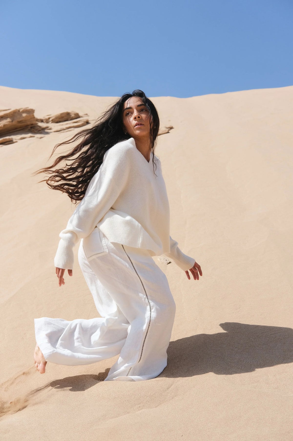 Woman in a white outfit standing on sand dunes with a clear blue sky.