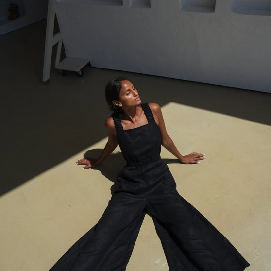 Woman in a black linen jumpsuit sitting on a light-colored floor with a white wall in the background
