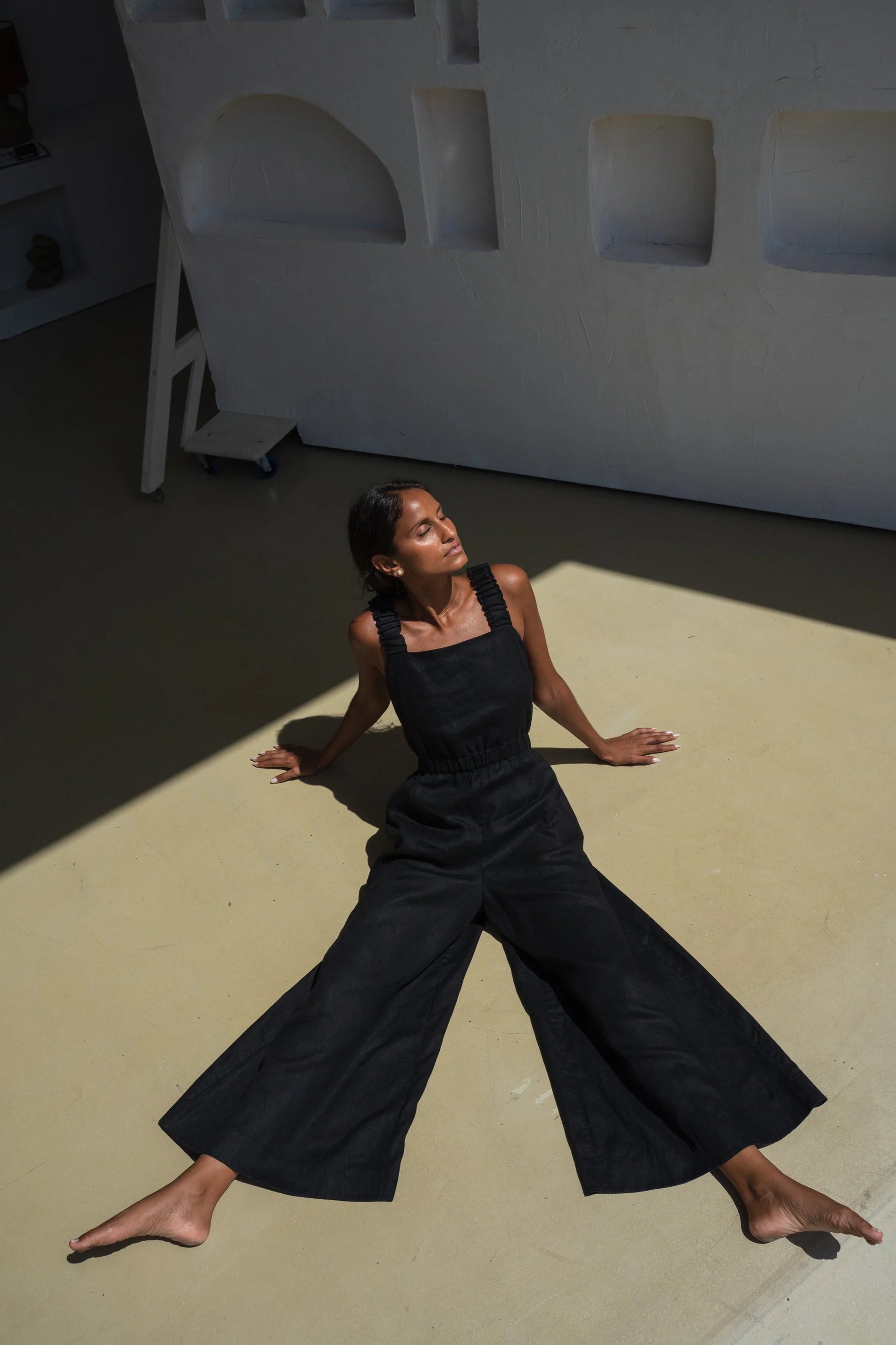 Woman in a black linen jumpsuit sitting on a light-colored floor with a white wall in the background