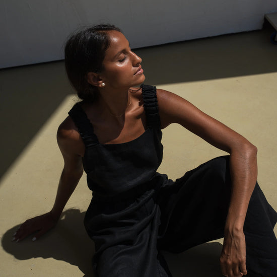 Woman in a black linen jumpsuit sitting on the floor with a minimal background
