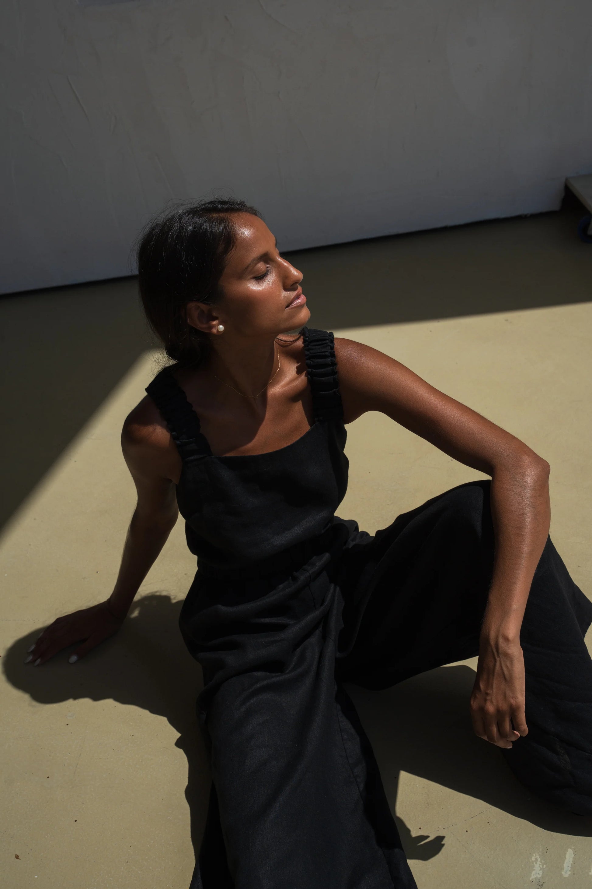 Woman in a black linen jumpsuit sitting on the floor with a minimal background