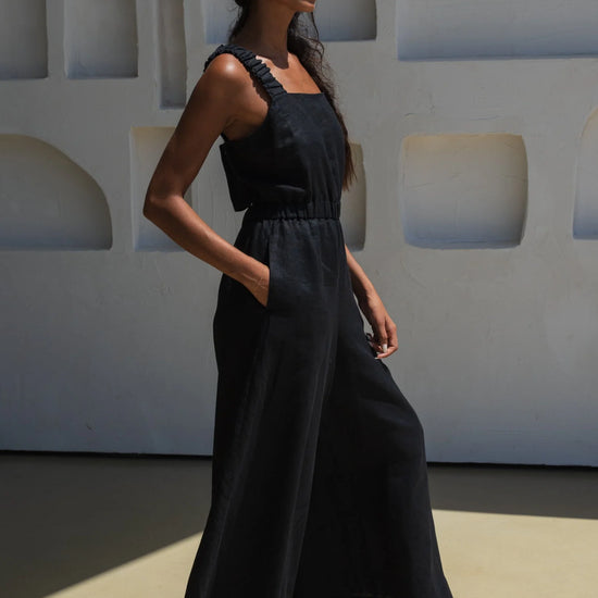 Woman in a black linen jumpsuit standing against a white wall with geometric shapes.