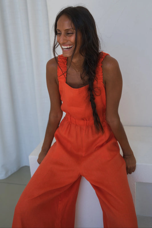 Woman in an orange linen jumpsuit sitting on a white bench with a white curtain background