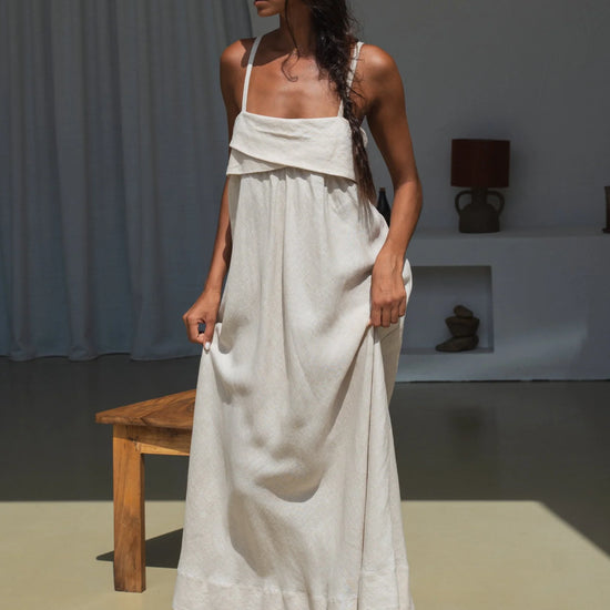 Woman in a beige linen dress standing in a minimalistic room with a wooden bench and lamp.