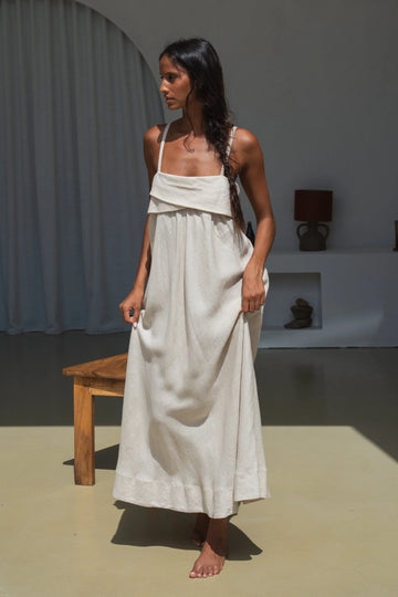 Woman in a beige linen dress standing in a minimalistic room with a wooden bench and lamp.