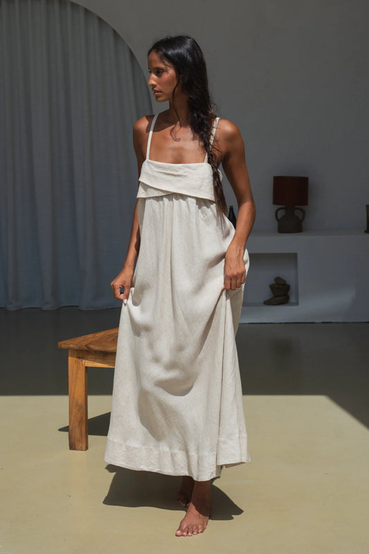Woman in a beige linen dress standing in a minimalistic room with a wooden bench and lamp.