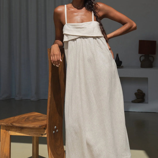 Woman in a long beige linen dress holding a wooden object in a minimalistic room.