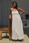 Woman in a long beige linen dress holding a wooden object in a minimalistic room.