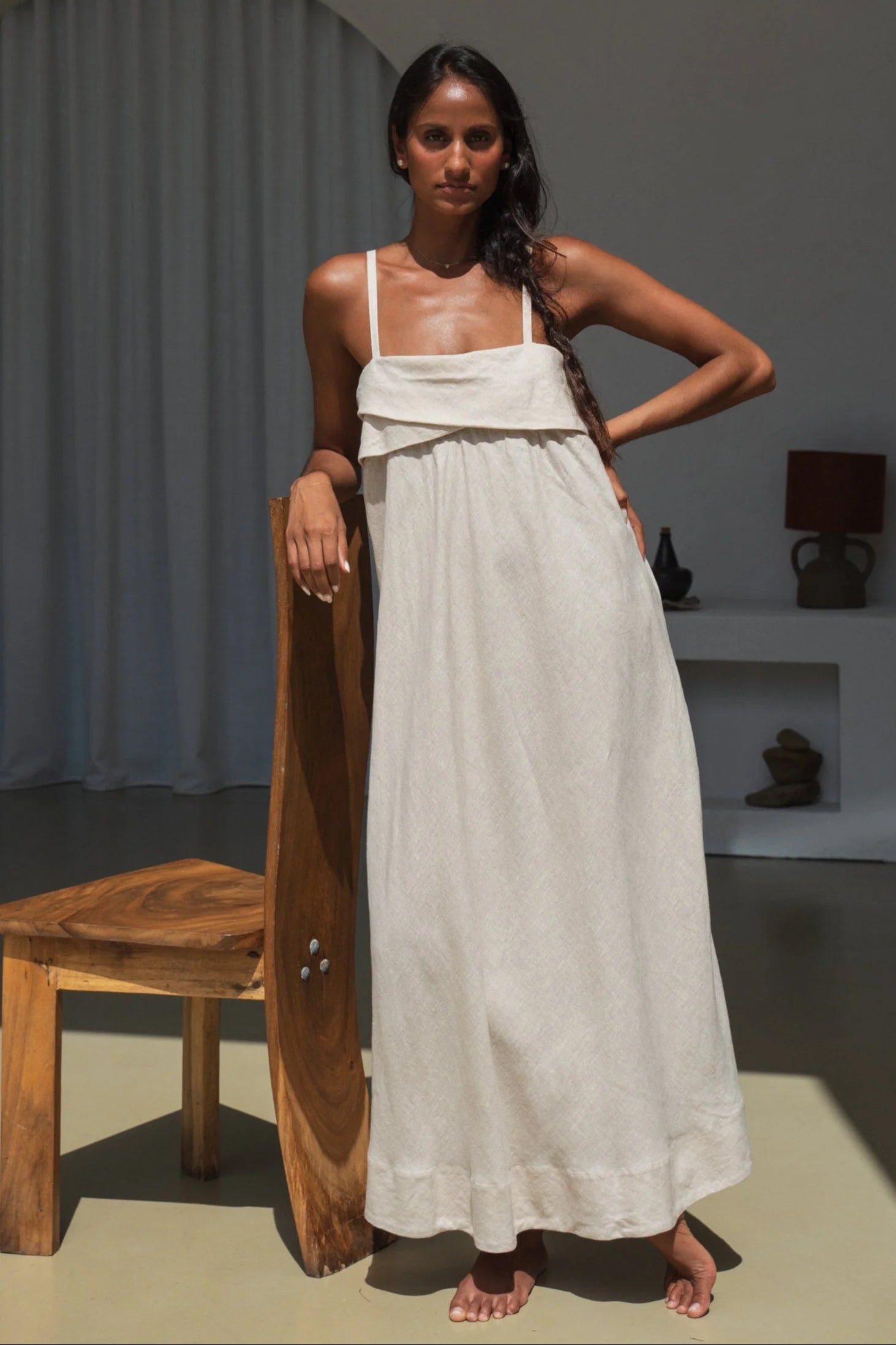 Woman in a long beige linen dress holding a wooden object in a minimalistic room.