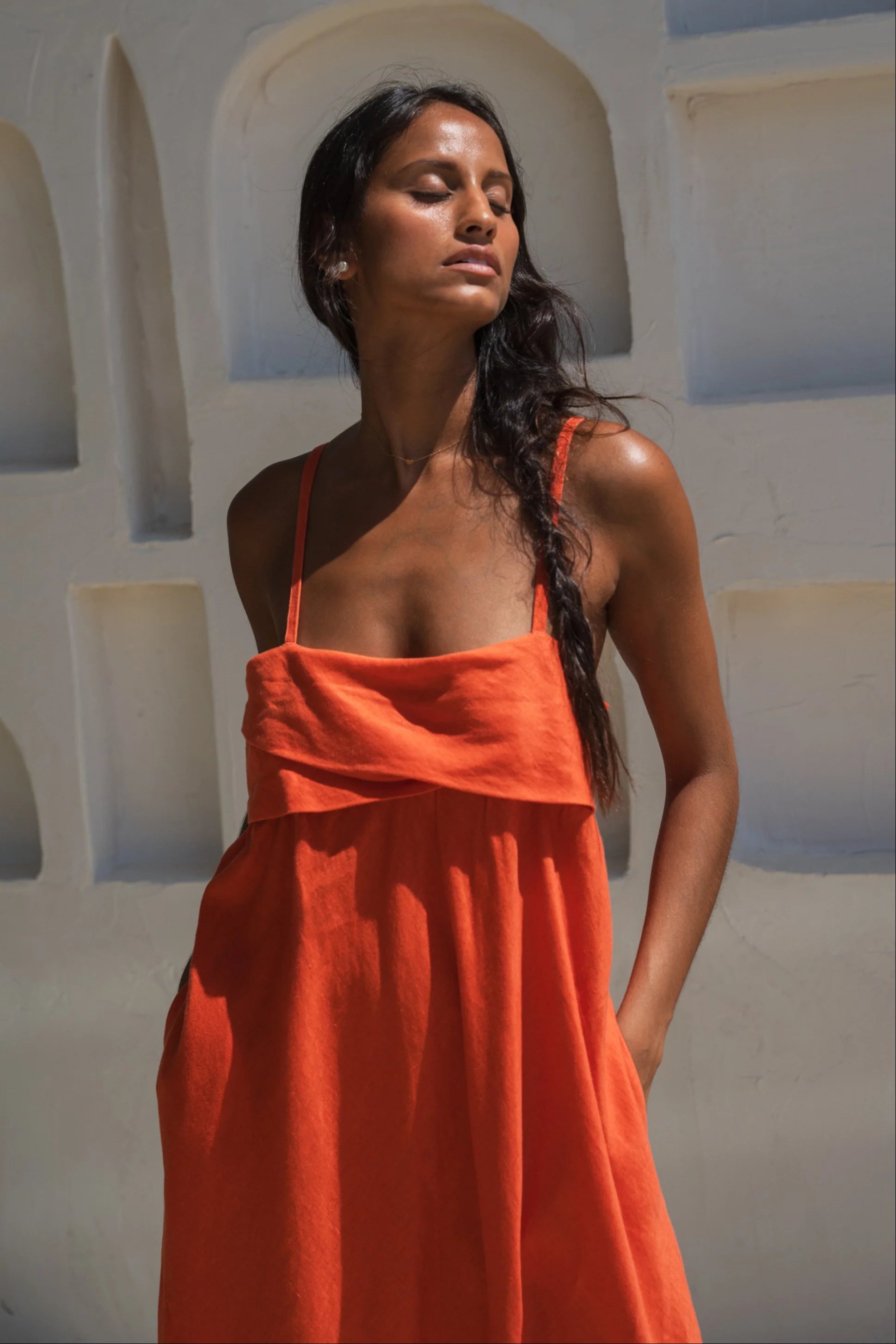 Woman wearing an orange linen dress standing against a white wall with arches.