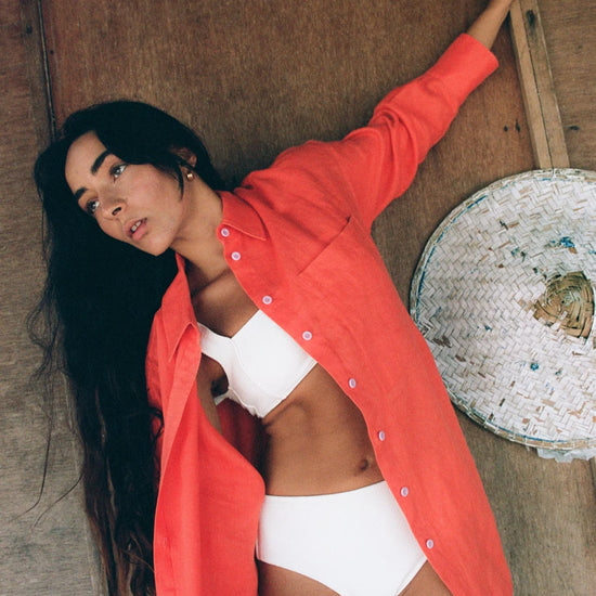 Woman wearing a bright orange shirt over a white bikini, standing against a textured wall.