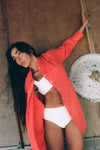 Woman wearing a bright orange shirt over a white bikini, standing against a textured wall.