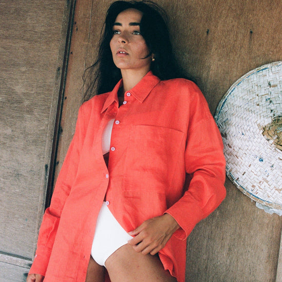 Woman wearing a bright orange shirt from lilly pilly over a white swimsuit, leaning against a wooden wall.