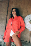 Woman wearing a bright orange shirt from lilly pilly over a white swimsuit, leaning against a wooden wall.