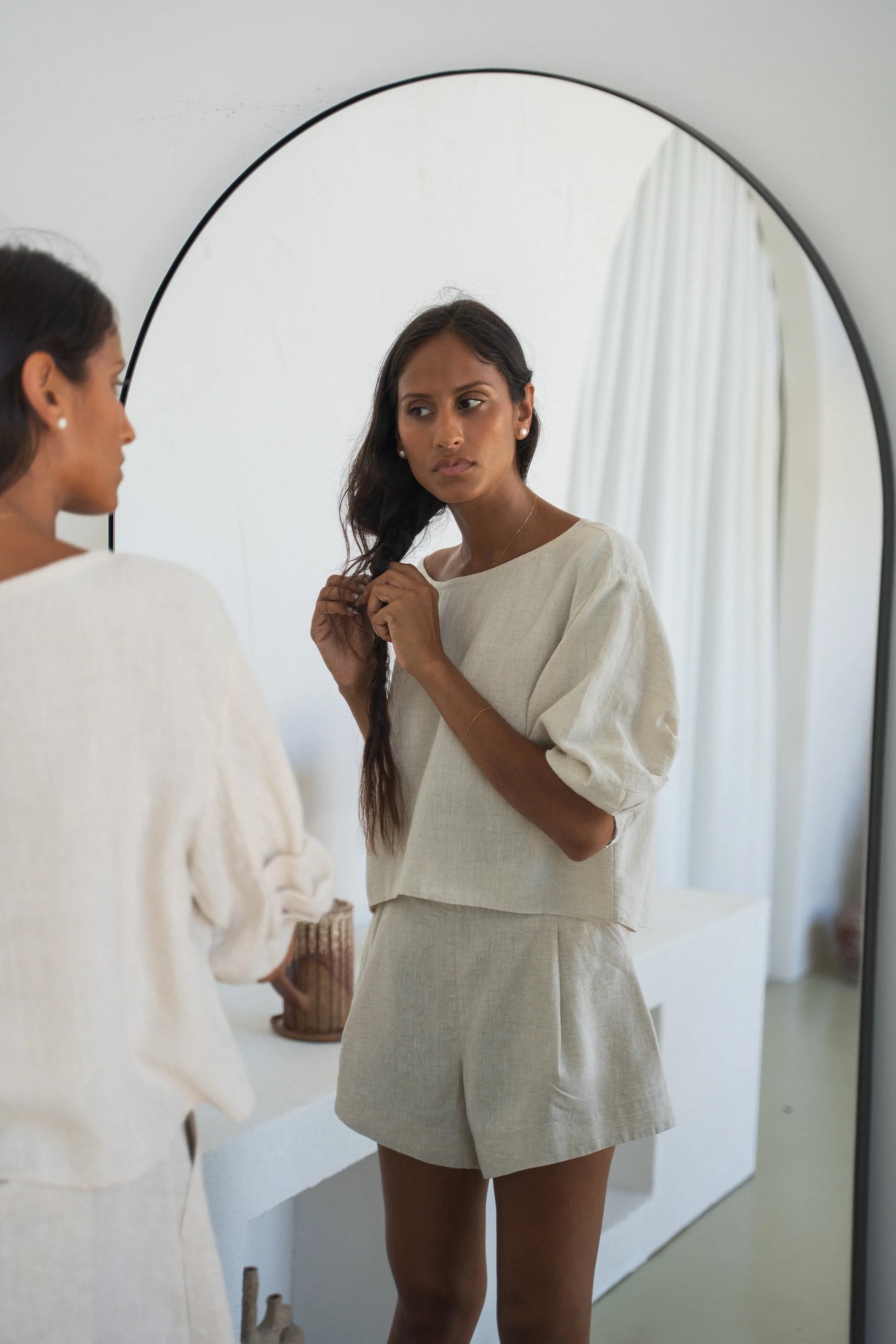 Woman adjusting her hair in front of a mirror wearing a light-colored outfit from Lilly Pilly