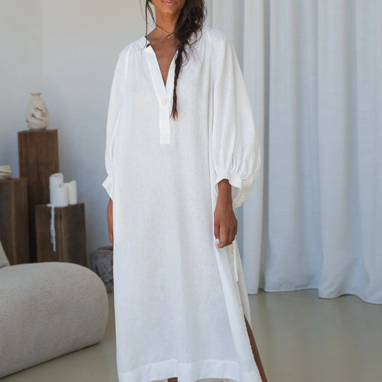 Woman wearing a white linen dress standing in a minimalistic room with white curtains and a light-colored floor.