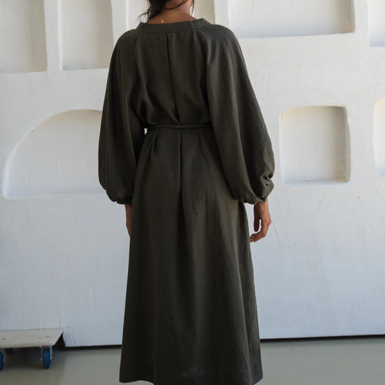Person wearing a long dark green linen dress standing in a minimalistic room with white walls and floor.