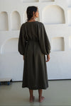 Person wearing a long dark green linen dress standing in a minimalistic room with white walls and floor.