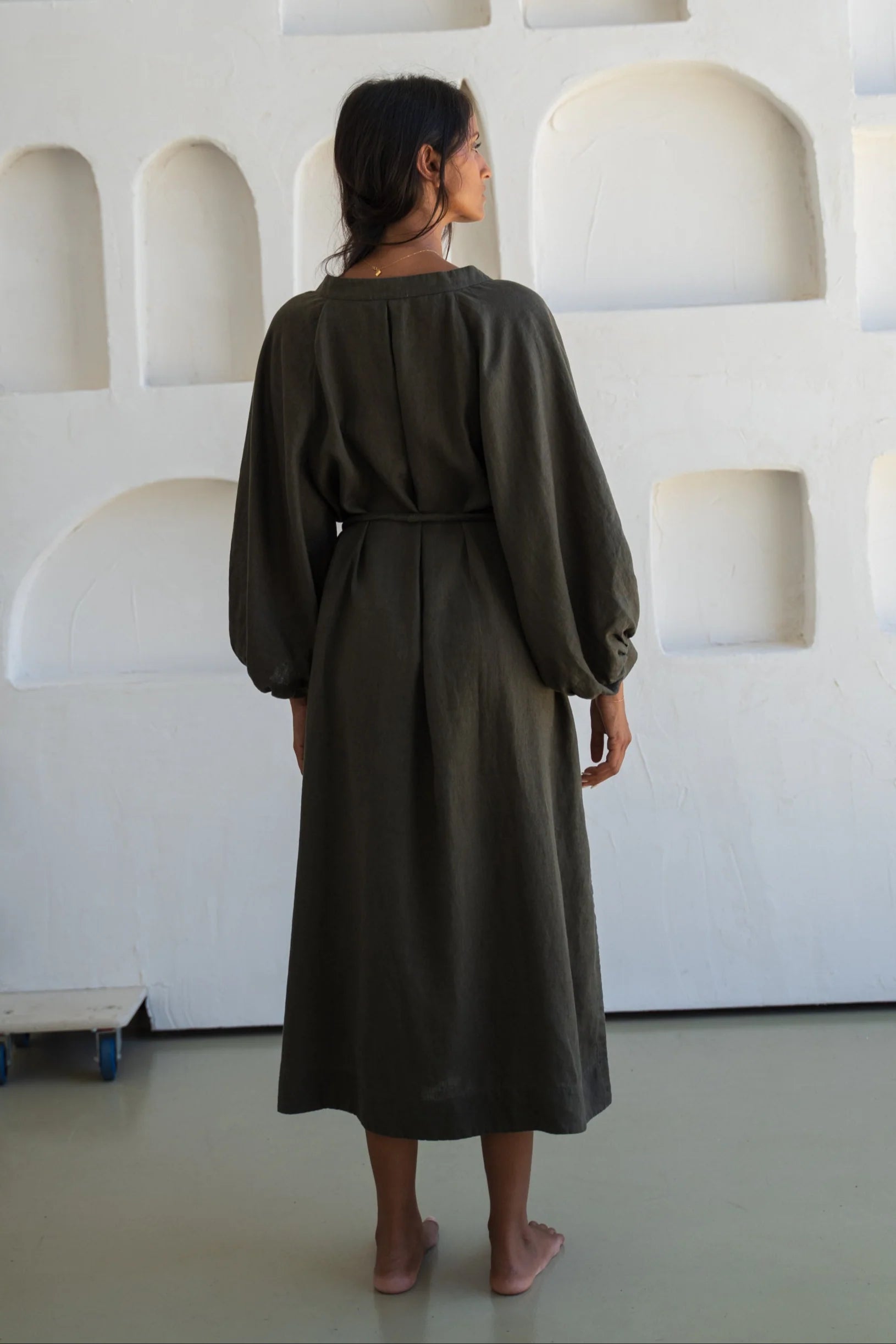 Person wearing a long dark green linen dress standing in a minimalistic room with white walls and floor.