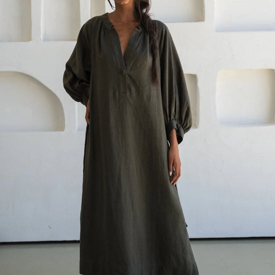 Woman wearing a long olive green linen dress standing against a white wall with geometric shapes.