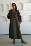 Woman wearing a long olive green linen dress standing against a white wall with geometric shapes.