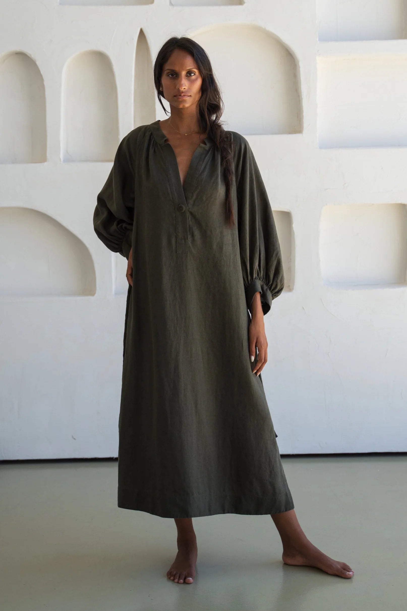 Woman wearing a long olive green linen dress standing against a white wall with geometric shapes.