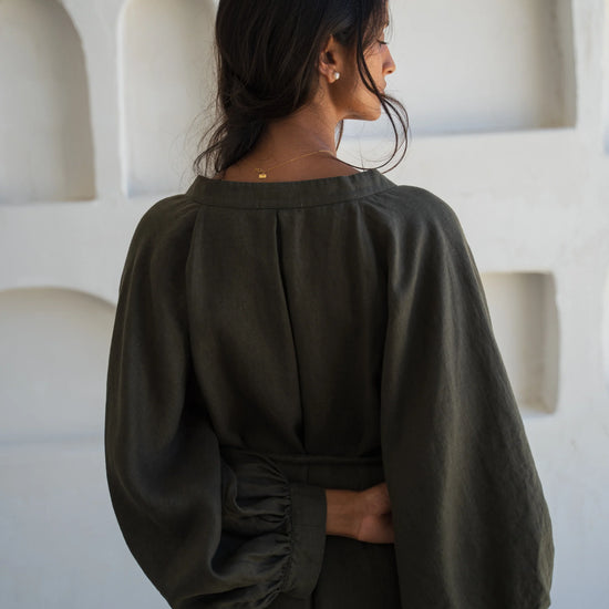 Woman wearing a dark green linen dress standing against a white wall with geometric shapes.