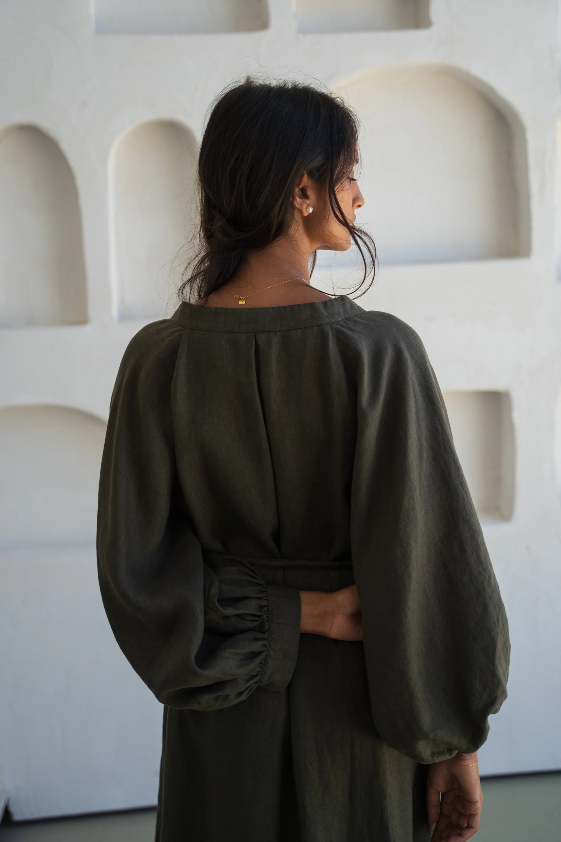 Woman wearing a dark green linen dress standing against a white wall with geometric shapes.