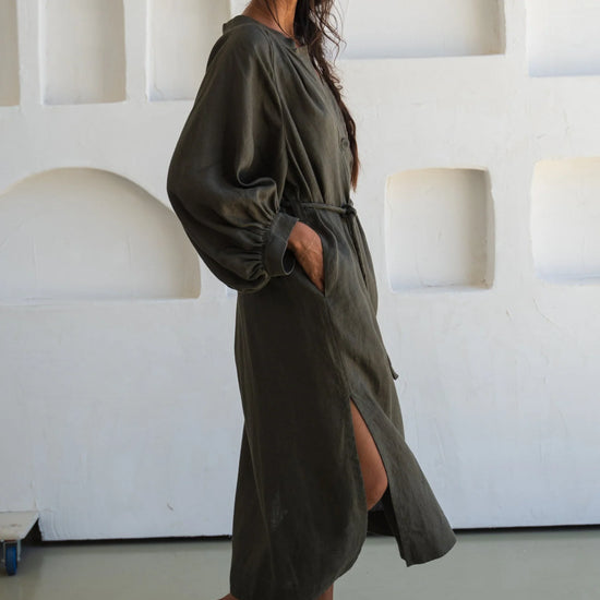 Woman wearing a long olive green linen dress standing against a white textured wall.
