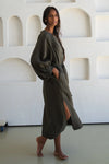 Woman wearing a long olive green linen dress standing against a white textured wall.