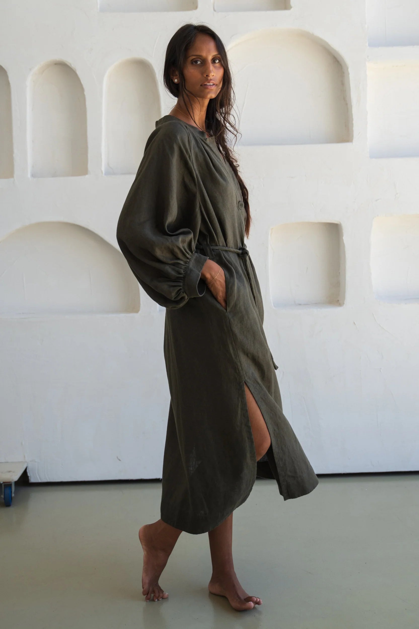 Woman wearing a long olive green linen dress standing against a white textured wall.