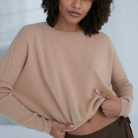 Woman wearing a beige sweater and brown pants against a neutral background