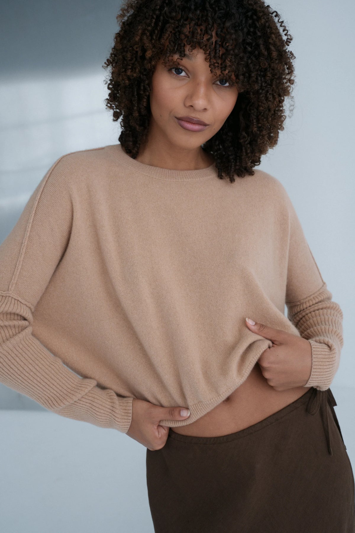 Woman wearing a beige sweater and brown pants against a neutral background