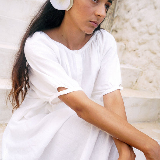 Woman in a white dress with a flower in her hair, sitting against a stone wall.
