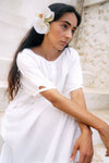 Woman in a white dress with a flower in her hair, sitting against a stone wall.