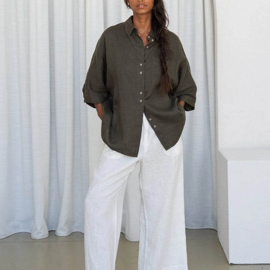 Person wearing a khaki linen shirt and white pants standing against a white curtain.