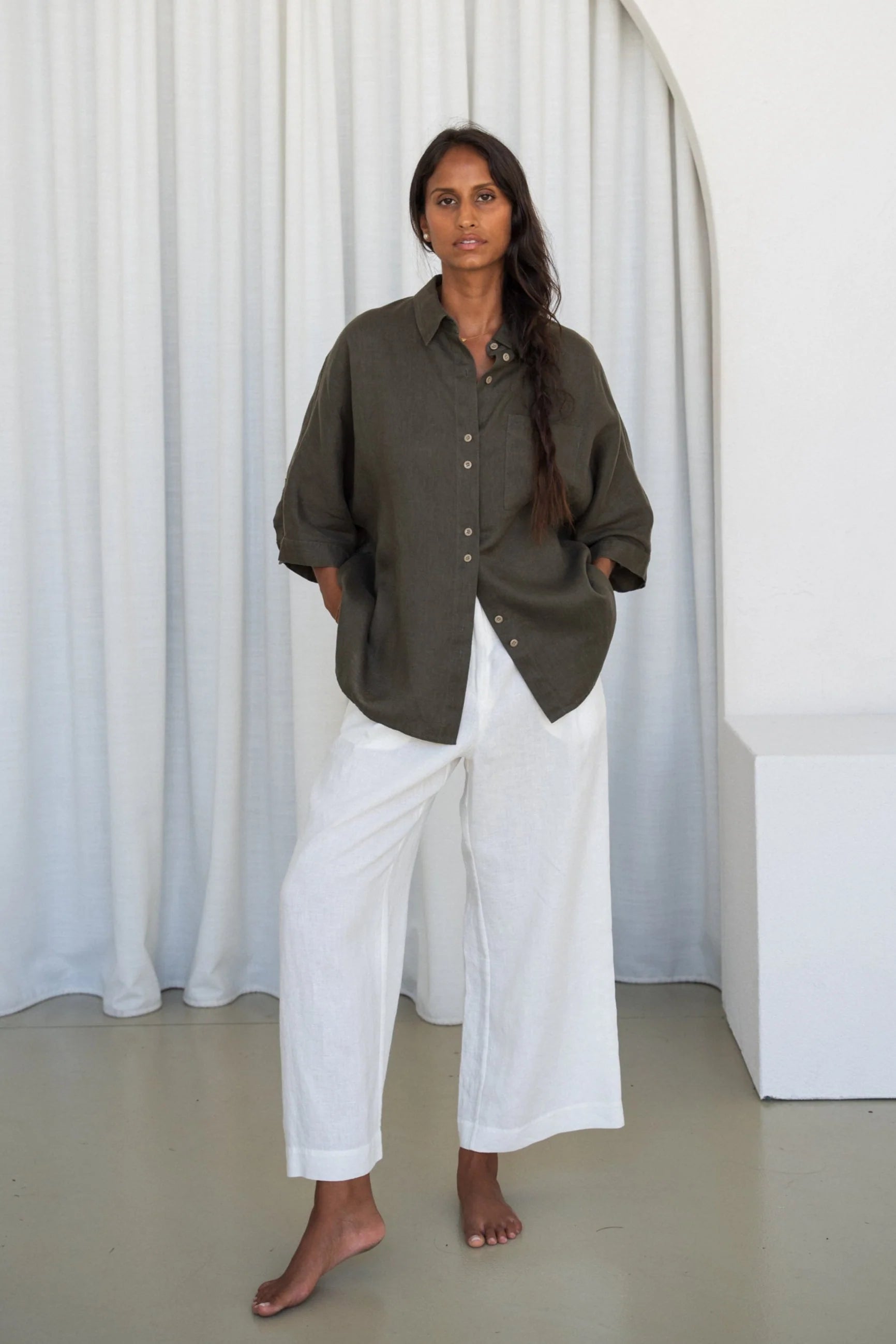 Person wearing a khaki linen shirt and white pants standing against a white curtain.