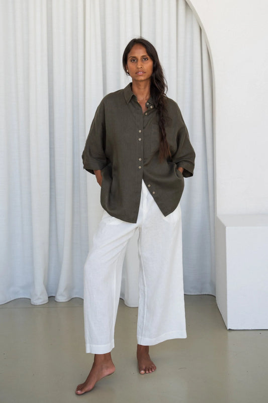 Person wearing a khaki linen shirt and white pants standing against a white curtain.