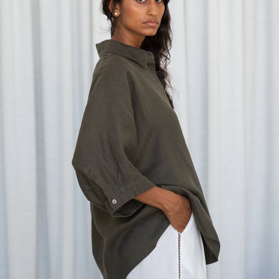 Woman wearing an okhaki green linen shirt and white pants against a white curtain background