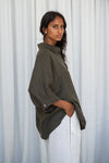 Woman wearing an okhaki green linen shirt and white pants against a white curtain background