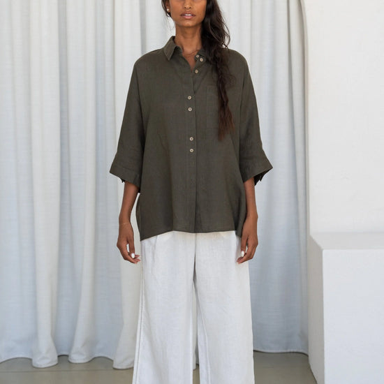 Woman wearing a khaki shirt and white pants standing against a white curtain.