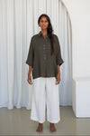 Woman wearing a khaki shirt and white pants standing against a white curtain.