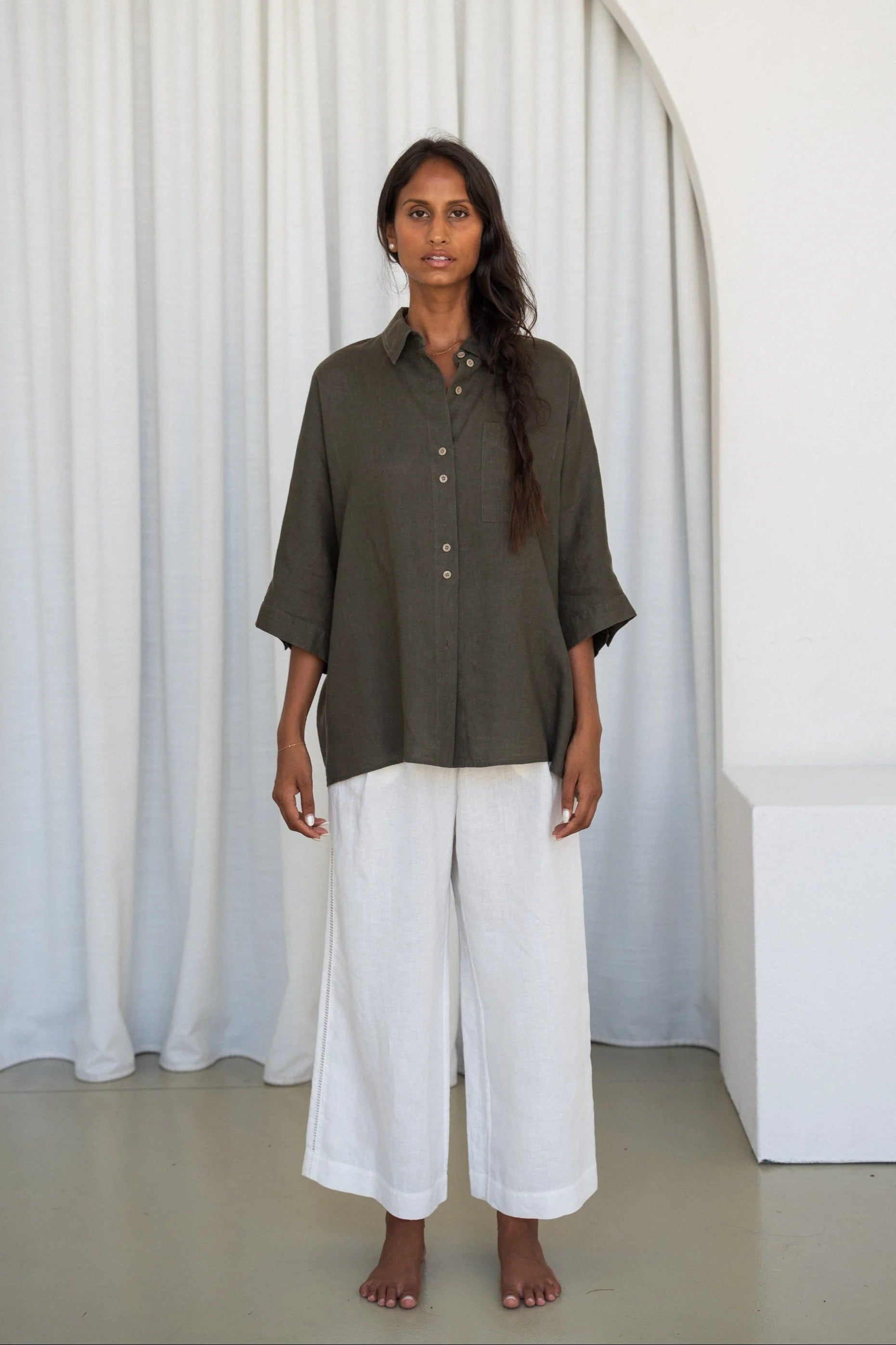 Woman wearing a khaki shirt and white pants standing against a white curtain.